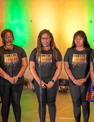 Un groupe de six artistes noires debout sur scène avec un rétroéclairage coloré. Visibilité masquée.
