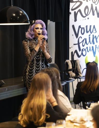 Une artiste de drag aux cheveux violets et à la robe argentée et noire s'exprime devant un groupe qui brunch. Visibilité masquée.