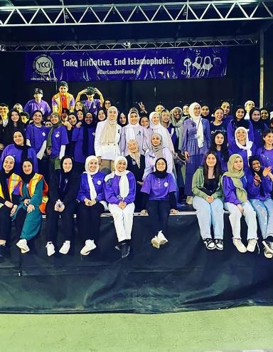 Des dizaines de personnes sont rassemblées sur une scène, la plupart souriantes et vêtues de violet. Beaucoup portent le hidjab. Une banderole est suspendue à l’arrière de la scène sur laquelle on peut lire, en anglais « Prenons l’initiative. Mettons fin à l’islamophobie. #OurLondonFamily ». Visibilité masquée.