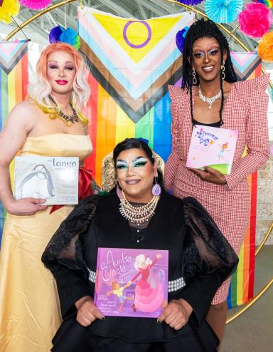 Three drag artists holding books are standing in front of a rainbow-themed circle arch. Visibilité masquée.