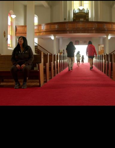 Intérieur d’une église avec une longue allée recouverte d’un tapis rouge entre des bancs en bois. La lumière du soleil filtre à travers les vitraux tandis que plusieurs enfants courent vers l’entrée lumineuse au fond de l’église et qu’une femme autochtone est assise seule sur un banc à gauche. Visibilité masquée.