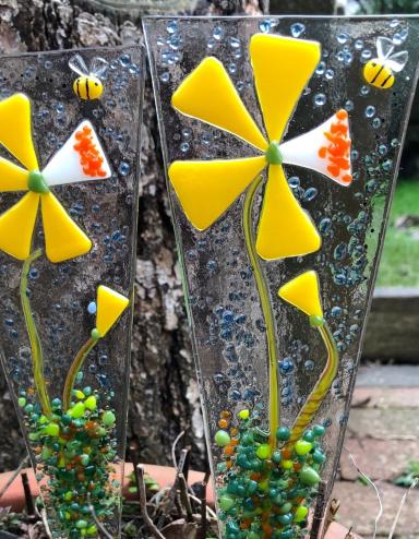 Deux décorations en verre fusionné représentant des fleurs jaunes sont placées dans un pot à fleurs à l’extérieur. Visibilité masquée.
