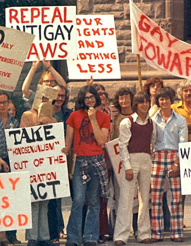 Un grand groupe de personnes vêtues de t-shirts et de pantalons évasés à la mode des années 1970 sont debout ensemble et tiennent des pancartes portant des messages en anglais tels que « sortis du placard », « c’est bien d’être gai » et « abrogez les lois anti-gaies ». Visibilité masquée.