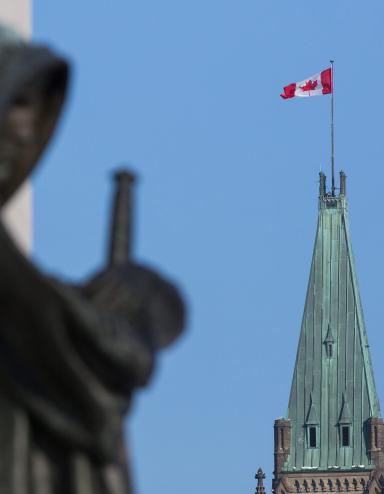 Un drapeau canadien flotte au sommet d’une grande tour. Au premier plan se dresse la statue d’une femme voilée portant une épée. Visibilité masquée.