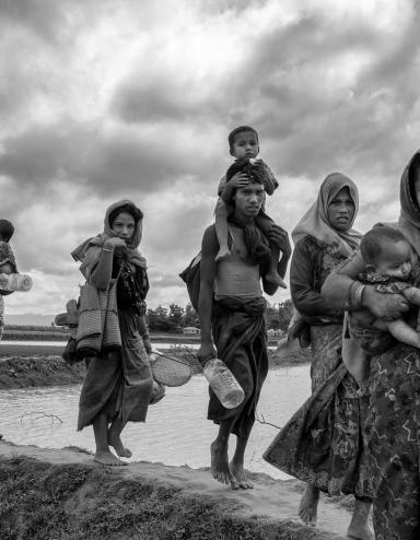 Un groupe de femmes et d’hommes rohingyas transportent de jeunes enfants et des effets personnels en marchant sur une digue de terre qui traverse une étendue d’eau. Visibilité masquée.
