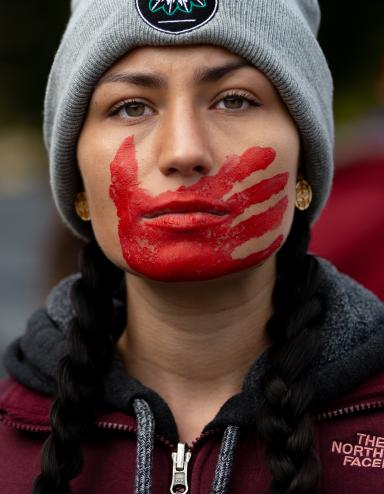 Une jeune femme aux cheveux tressés, vêtue d’une veste bordeaux et coiffée d’un bonnet d’hiver gris, regarde résolument vers l’avant, une empreinte de main rouge peinte sur la bouche et le visage. Visibilité masquée.