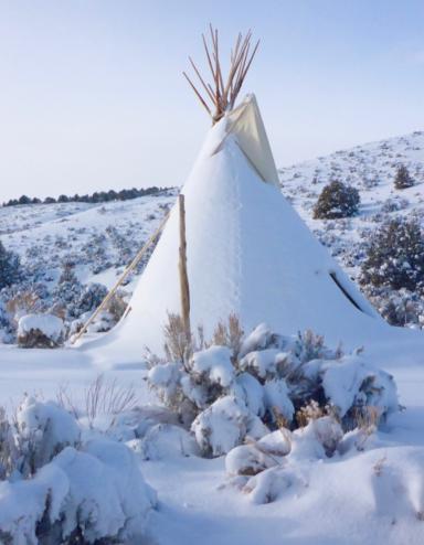 Un tipi recouvert de neige se dresse dans un paysage vallonné parsemé d’arbres et de buissons, sous un ciel bleu. Visibilité masquée.