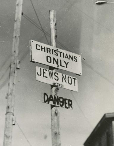 Photo noir et blanc de trois écriteaux fixés sur un poteau, où l’on peut lire en anglais « Chrétiens seulement », « Interdit aux Juifs » et « Danger ». Visibilité masquée.