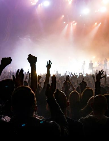 Des personnes qui assistent à un concert lèvent les mains en signe de jubilation collective tandis qu’un groupe joue sur une scène brillamment éclairée. Visibilité masquée.