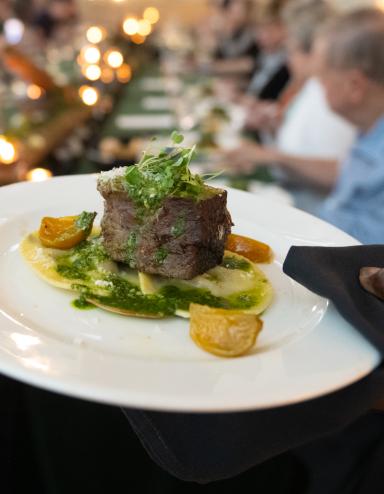Au premier plan, la main d'un serveur tenant une assiette de steak avec des garnitures est nette, tandis qu'une table de convives est floue à l'arrière-plan. Visibilité masquée.
