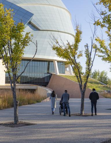 Quatre personnes suivent le chemin menant au Musée canadien pour les droits de la personne par une journée d'automne. Trois d'entre elles sont à pied et une personne est en fauteuil roulant. Visibilité masquée.