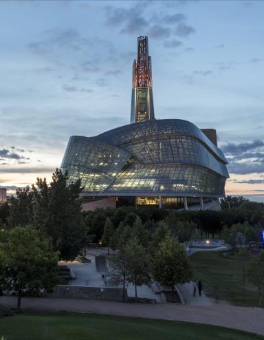 Photo extérieure des fenêtres en verre et de la tour du Musée canadien pour les droits de la personne, prise au crépuscule avec le ciel des prairies en arrière-plan. Le panneau Winnipeg à Forks est situé à droite de l'image. Plusieurs tours de bureaux et les lumières du centre-ville sont visibles à gauche de l'image. Visibilité masquée.