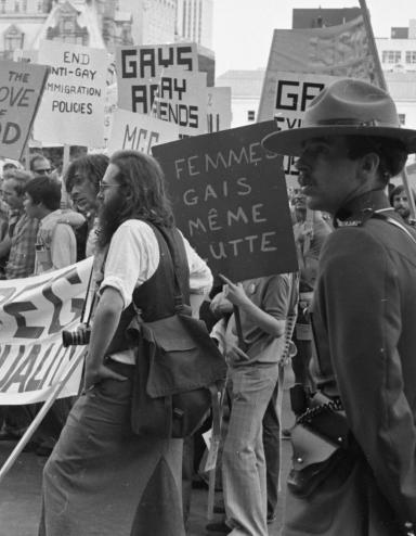 Un grand groupe de protestataires défile dans une ville tandis que deux agents de la GRC en uniforme surveillent la scène. Sur cette image en noir et blanc, les pancartes et les banderoles réclament la fin des pratiques discriminatoires contre les membres de la communauté 2ELGBTQI+. Visibilité masquée.