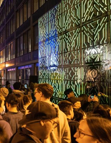 Beaucoup de gens se tiennent entre deux miroirs joints aux motifs complexes qui reflètent les lumières vertes et une foule de spectateurs. Visibilité masquée.