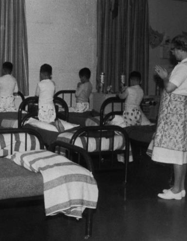 Un groupe de garçons en pyjama est agenouillé sur des lits simples, la tête baissée et les mains jointes comme pour prier. Une femme se tient dans la pièce, les mains jointes de la même manière. Visibilité masquée.