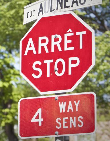 Un panneau d’arrêt et un panneau de signalisation de rue, en français et en anglais, se trouvent devant des arbres. Visibilité masquée.