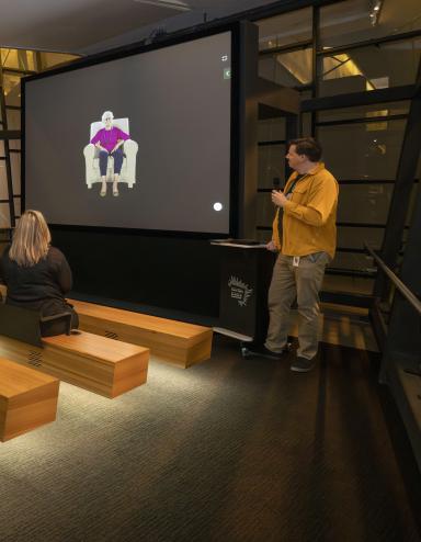 Un petit groupe de personnes assises sur des bancs en bois dans un théâtre fait face à un écran montrant une femme âgée assise dans un fauteuil. Un homme vêtu d’une chemise jaune se tient à droite de l’écran et tient un microphone. Visibilité masquée.