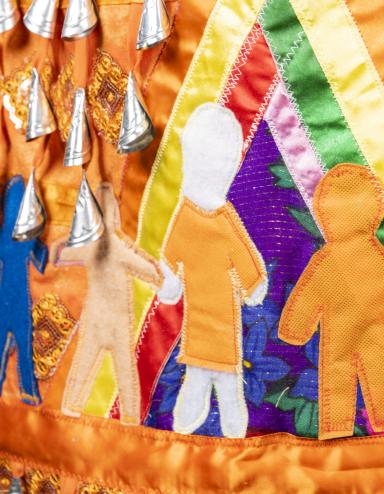 Une section d'une robe orange à clochettes est représentée. Les clochettes, ou petits pendentifs argentés en forme de cône, sont visibles en haut de l'image. Sous elles, sept figures humaines sont représentées, toutes de couleurs et de tailles différentes. L'une des figures porte une chemise orange. Ces figures humaines se tiennent la main à l'intérieur d'un tipi multicolore.