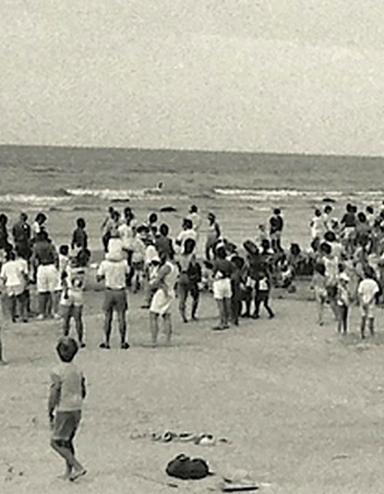 Une photographie en noir et blanc d’une foule de personnes, la plupart debout, sur une plage.