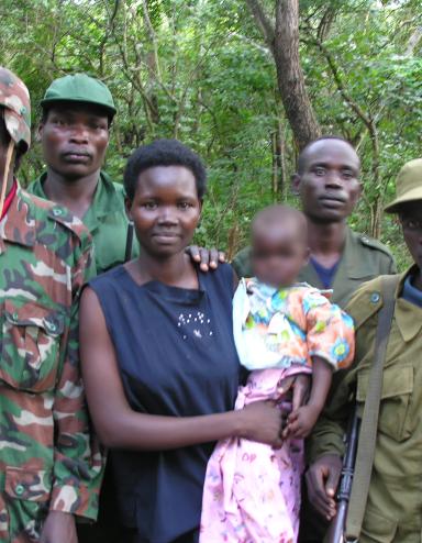 Une femme tenant un bébé, tandis que quatre hommes en tenue militaire se tiennent à côté et derrière elle. Ils sont tous debout devant une forêt et posent pour la photo.