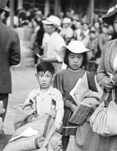 Photo en noir et blanc d’une femme avec deux enfants qui se tiennent derrière une pile de bagages et de couvertures et qui fixent l’objectif.