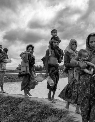 Un groupe de femmes et d’hommes rohingyas transportent de jeunes enfants et des effets personnels en marchant sur une digue de terre qui traverse une étendue d’eau.