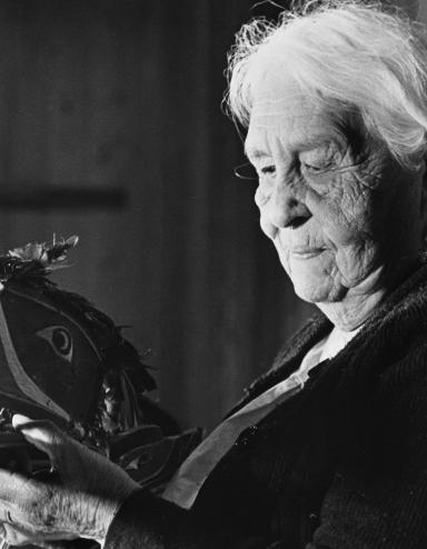 Une image en noir et blanc d’une femme âgée aux cheveux blancs tenant un masque formé et peint de façon à représenter la tête d’un oiseau. Le long bec est ouvert, et il y a des plumes attachées sur le haut du masque. La femme regarde le masque qu’elle tient dans ses mains.