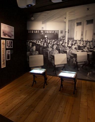Une exposition muséale montrant une photo en noir et blanc d’enfants assis à des pupitres placés en rangées. Deux pupitres semblables à ceux qu’on voit dans la photo sont placés au centre de l’exposition. Sur un panneau de texte, on peut lire « Enfances oubliées ». Visibilité masquée.