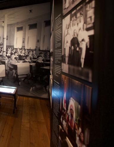 Une exposition muséale montrant une photo en noir et blanc d’enfants assis à des pupitres placés en rangées. Deux pupitres semblables à ceux qu’on voit dans la photo sont placés au centre de l’exposition. Sur un panneau de texte, on peut lire « Enfances oubliées ». Visibilité masquée.