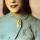 Portrait en buste d’une Viola Desmond souriante. Elle porte une veste bleu pâle à motifs brodés. La veste est fermée au col par une broche en forme de main avec deux doigts écartés pour former le V de la victoire.