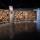 Une grande installation artistique horizontale à armature en bois composé de multiples losanges et rectangles sur lesquels sont fixés de nombreux objets. Une porte est entrouverte en plein centre de l’œuvre.