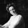 Photographie noir et blanc de Nellie McClung assise à un bureau. Elle porte un chemisier blanc et tient une plume. Sur le bureau se trouvent une lampe, un encrier et du papier.