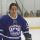 A young man in a blue hockey uniform with a crown logo on an indoor ice rink.