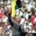 Nelson Mandela, un homme plus âgé à la peau foncée et aux cheveux gris et courts, est debout sur un podium et parle dans deux microphones. Il porte un complet gris et une cravate. Il lève son poing droit au-dessus de sa tête.