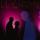 Les silhouettes d’un homme et d’une femme qui se font face et se tiennent devant un mur sombre éclairé de taches de lumière violette.