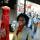 Une femme souriante se tient devant une robe de bal rouge et un smoking noir exposés sur des mannequins. La robe et le costume sont derrière une vitrine.