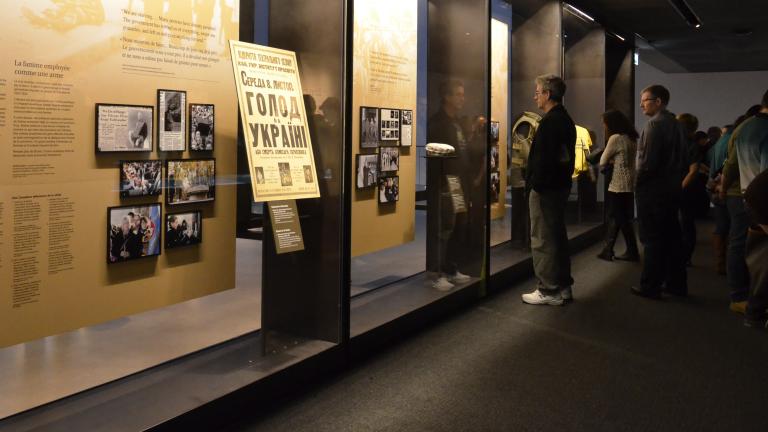 Un groupe de personnes regardent une série de vitrines qui vont du plancher au plafond. Les vitrines contiennent de grands panneaux de texte et de plus petits panneaux avec des images. Visibilité masquée.