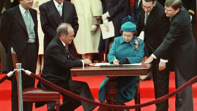 La reine Elizabeth II est assise à une table et signe un document. Un homme est assis à sa droite, et d’autres sont debout derrière eux autour de la table.