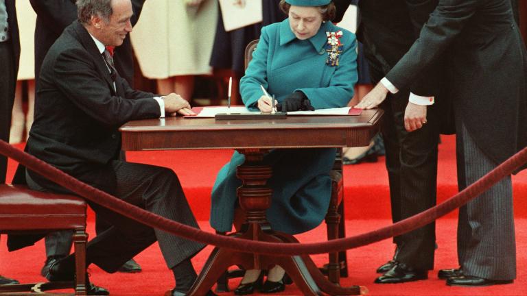 La reine Elizabeth II est assise à une table et signe un document. Un homme est assis à sa droite, et d’autres sont debout derrière eux autour de la table.