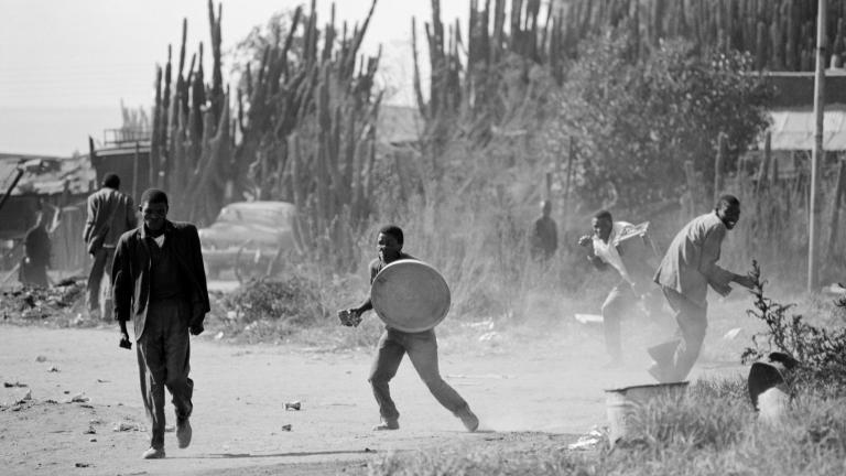 Photo en noir et blanc de quelques hommes noirs dans une environnement extérieur poussiéreux. L’un d’eux tient un couvercle de poubelle en guise de bouclier.