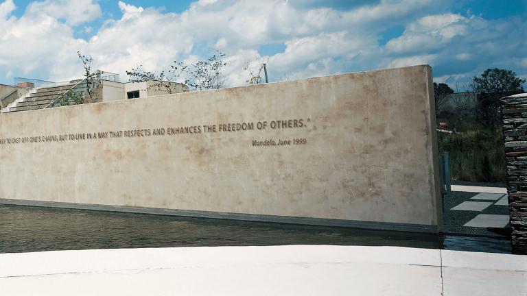 Un vue du Apartheid Museum. À l’avant-plan, on voit un plan d’eau. Dans l’eau se dresse un mur sur lequel on peut lire en anglais une citation de Nelson Mandela qui dit : « To be free is not merely to cast off one’s chains, but to live in a way that respects and enhances the freedom of others. »