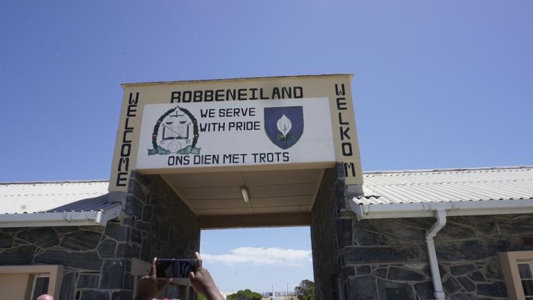 Une ouverture dans un bâtiment et un grand panneau au-dessus de l’ouverture. Sur panneau, on peut lire en anglais et en afrikaans « Welcome » et « We serve with pride ». Au haut du signe, on peut lire « Robbeniland ».