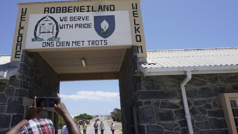 Une ouverture dans un bâtiment et un grand panneau au-dessus de l’ouverture. Sur panneau, on peut lire en anglais et en afrikaans « Welcome » et « We serve with pride ». Au haut du signe, on peut lire « Robbeniland ».