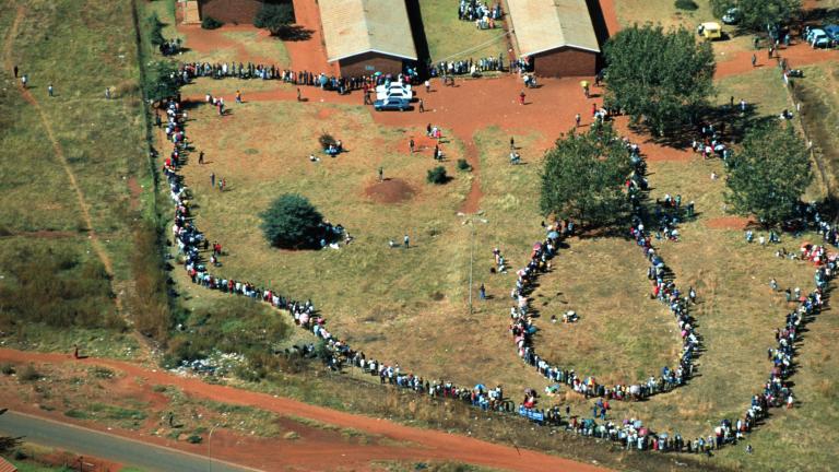 Vue aérienne d’une file de gens qui serpente à l’extérieur d’un grand bâtiment. La file est extrêmement longue, composée de centaines, voire de milliers de personnes.