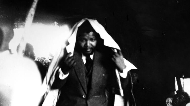 Une image en noir et blanc d’un jeune Nelson Mandela portant un complet, voûtant le dos et tenant un imperméable autour de sa tête, comme pour se protéger de la pluie.
