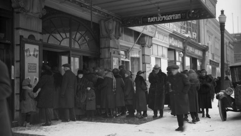 Une mage en noir et blanc d’une foule devant l’entrée d’un théâtre du nom de « Metropolitan ». Tout le monde porte de longs manteaux à l’ancienne, et certains portent des chapeaux. Une voiture antique est garée dans la rue.