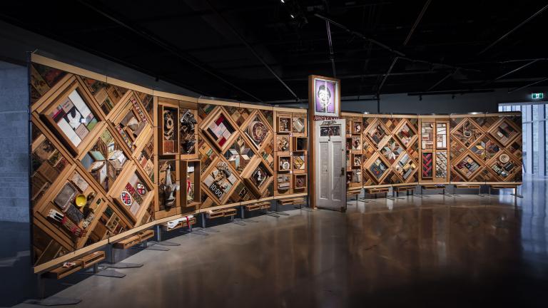 Une grande installation artistique horizontale à armature en bois composé de multiples losanges et rectangles sur lesquels sont fixés de nombreux objets. Une porte est entrouverte en plein centre de l’œuvre.