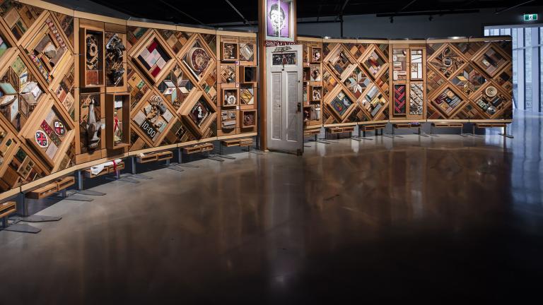 Une grande installation artistique horizontale à armature en bois composé de multiples losanges et rectangles sur lesquels sont fixés de nombreux objets. Une porte est entrouverte en plein centre de l’œuvre.