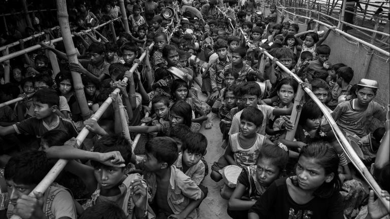 Des centaines d’enfants rohingyas, la plupart accroupis ou assis, font la queue pour de la nourriture. Les files d’attente sont séparées par de longues rangées de poteaux en bambou.