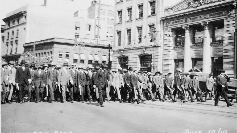 Un grand groupe d’hommes en costumes et chapeaux brandissant des matraques et marchant dans la rue.